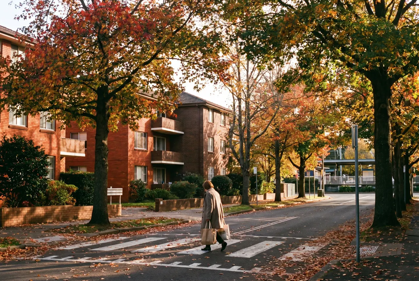 Low-rise brick apartment buildings among mature autumn trees near Gordon village, a resident crossing a leafy street